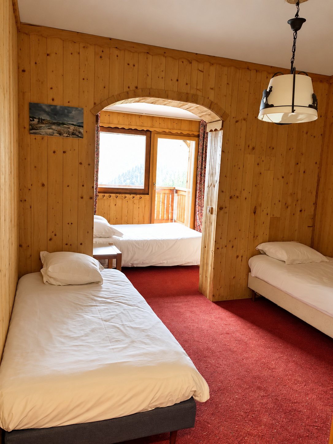 Chambre lambrissée de style chalet avec trois lits simples, moquette rouge et une alcôve voûtée donnant sur un balcon lumineux.
