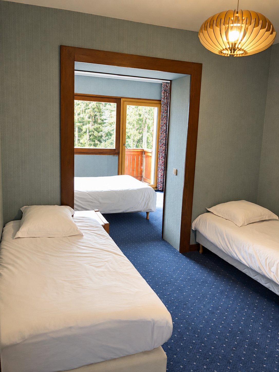 Chambre d'hôtel aux murs gris-vert avec trois lits aux draps clairs, moquette bleue, suspension en bois et porte-fenêtre ouvrant sur un balcon en bois.