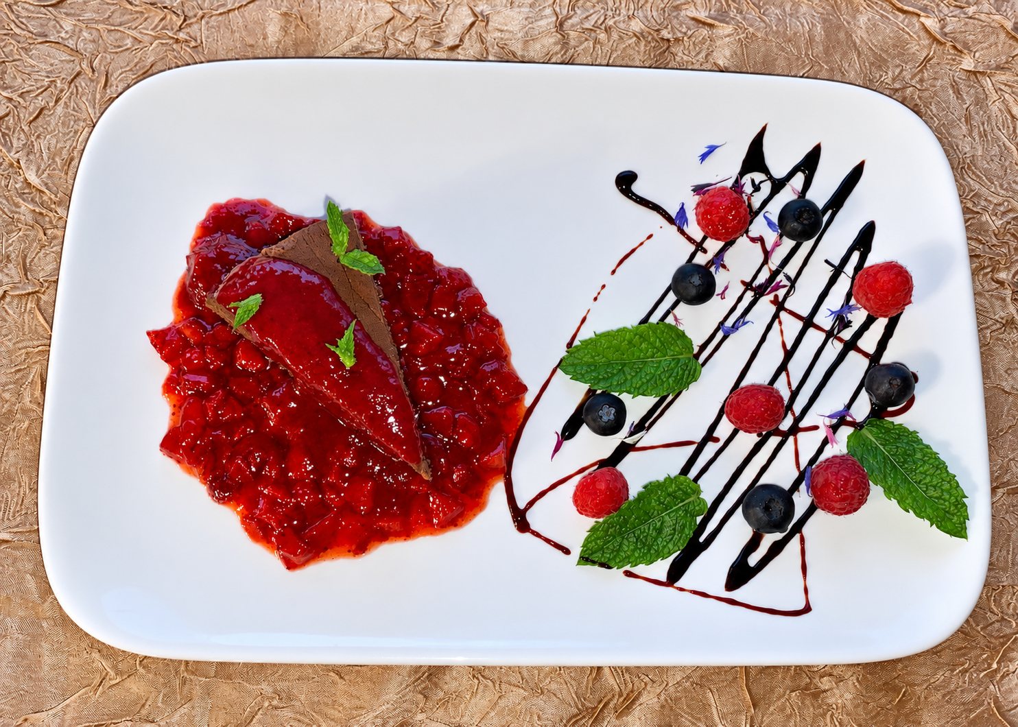 Assiette rectangulaire blanche avec une part de dessert au chocolat sur une compotée de fruits rouges, décorée de framboises, myrtilles, feuilles de menthe et d'un trait de coulis sombre.