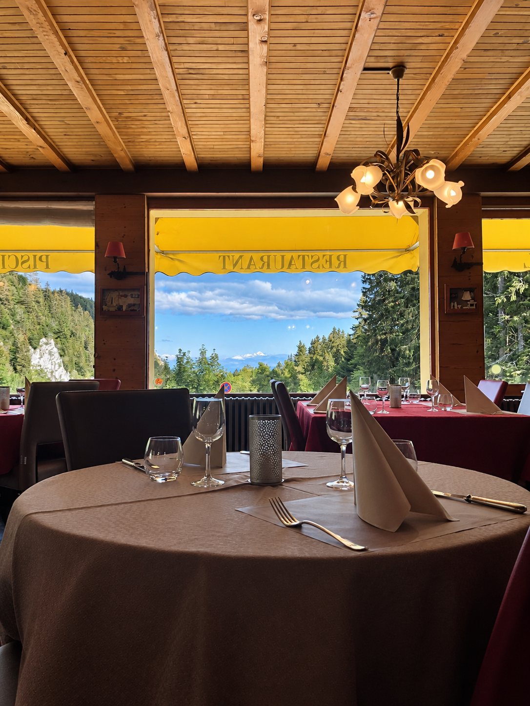 Table ronde dressée dans une salle de restaurant au plafond de bois, devant une grande baie vitrée donnant sur des sapins et le ciel.
