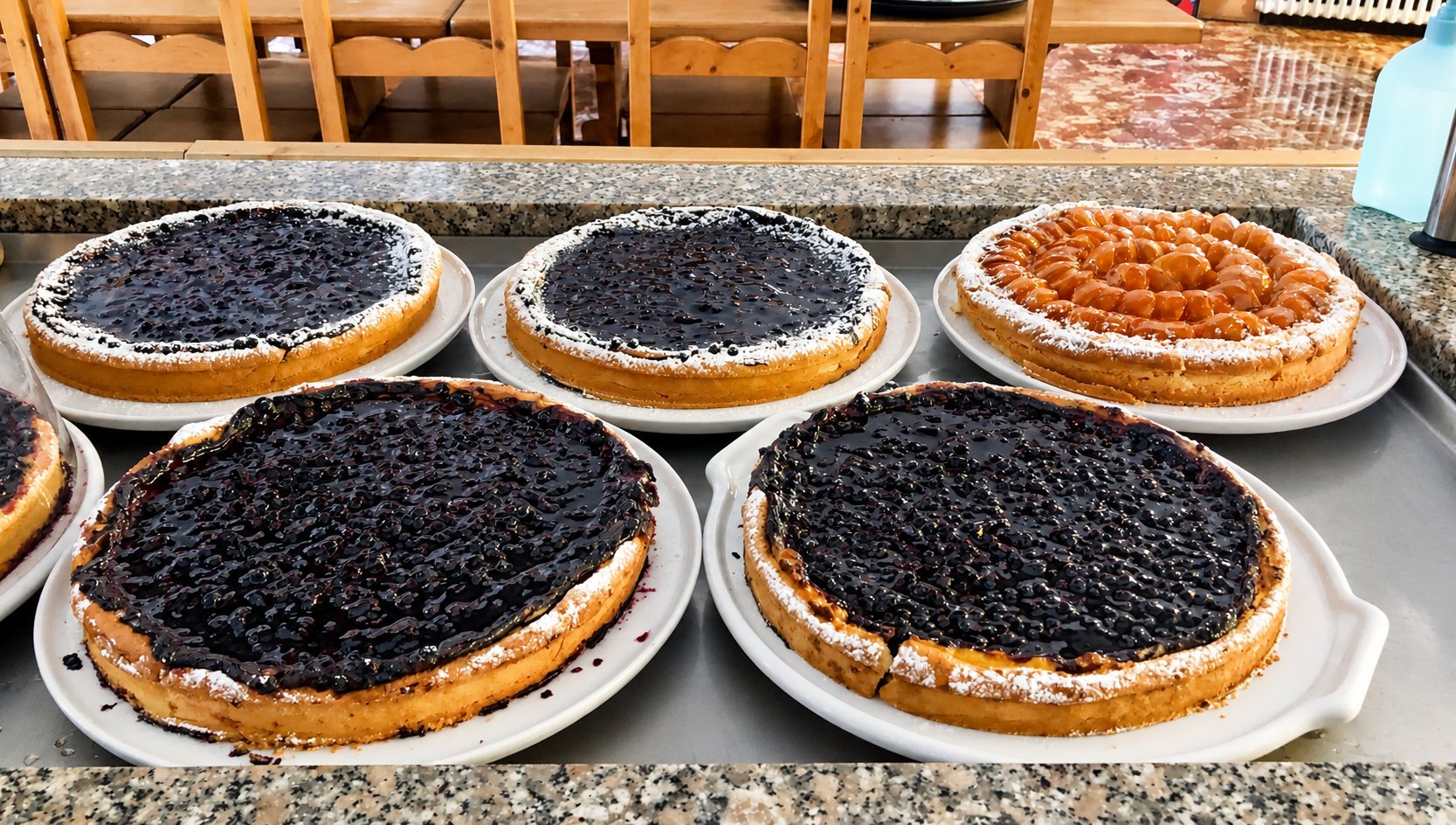 Cinq tartes maison, dont quatre aux myrtilles et une aux abricots saupoudrée de sucre glace, posées sur des assiettes blanches sur un comptoir en granit.